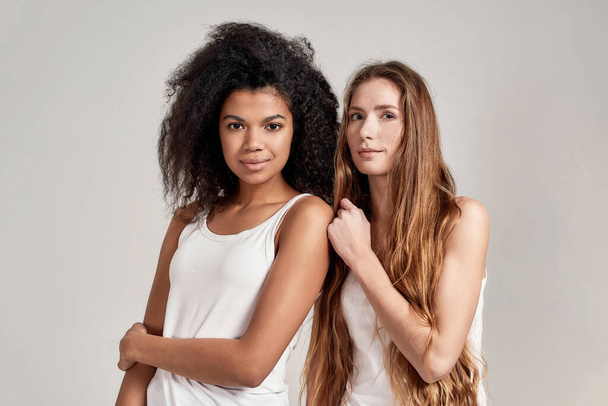 Portrait of two young diverse women wearing white shirts looking at camera while posing together isolated over grey background - Фото, изображение