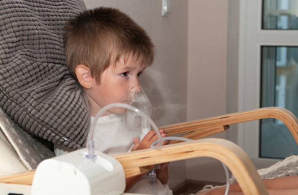 Small sick boy with nebulizer mask making inhalation, respiratory procedure by pneumonia or cough for child in home. - 写真・画像