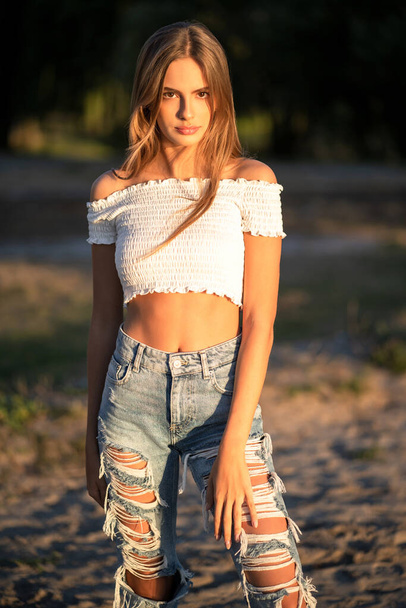 Hipster teenage girl looks at camera while posing on sunset outdoors. Fashion young model walks at beach in sunny evening - Foto, imagen