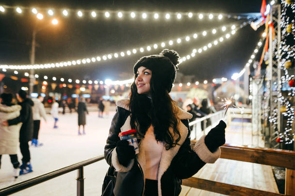 Egy vidám, meleg ruhás lány áll a jégpálya mellett egy csésze forró itallal a kezében, félrenéz, mosolyog és kezében egy világító csillagszórót tart. A hölgy az utcán ünnepli a karácsonyt.. - Fotó, kép