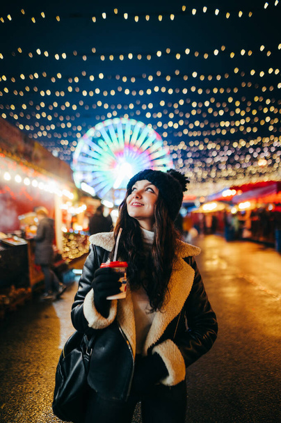 Šťastná dívka s šálkem horkého nápoje v ruce stojí večer na Vánoční korunované ulici, zvedá hlavu a vypadá a světlušky scenérie s úsměvem na tváři. Svisle. - Fotografie, Obrázek