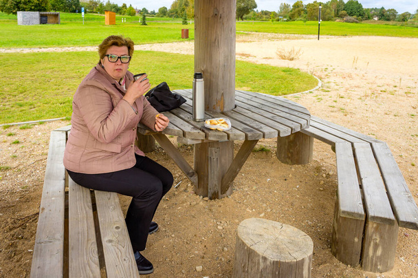 Mulher madura alegre descansando em um parque da cidade. Ela bebe chá com um pão - Foto, Imagem
