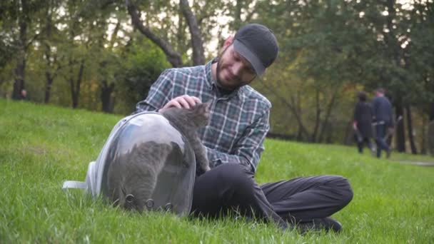 El hombre usa la mochila con ojo de buey para mascotas, sentado sobre hierba verde en el parque jugando y divirtiéndose con gato gris. El equipo es transparente respirar cápsula de la que divertido gato tira de la pata hacia el propietario - Imágenes, Vídeo