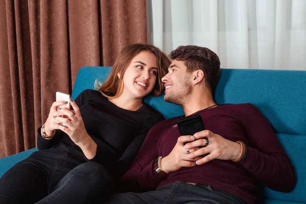 Photo of positive excited people man and woman, couple in love looking at each other smiling while both using mobile phones sitting, lying down on a blue sofa couch. - Foto, imagen