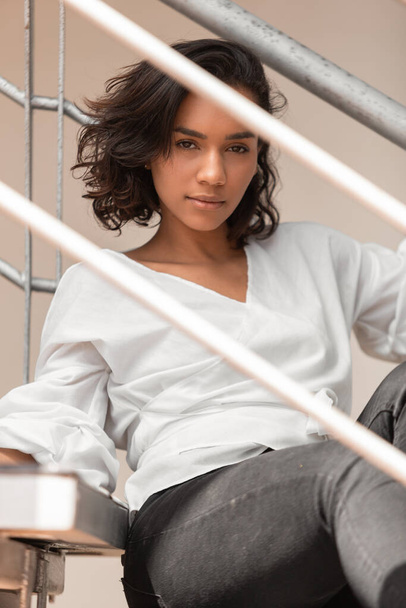 aantrekkelijke jonge latino brunette vrouw met krullen en kort haar zitten op de trap, het dragen van witte blouse en zwarte broek, foto overdag - Foto, afbeelding