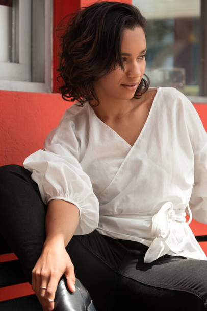 schattig jong Latijns brunette vrouw met krullen en kort haar, zitten op een bank buiten, het dragen van een witte blouse en zwarte broek, foto in de dag met zon - Foto, afbeelding