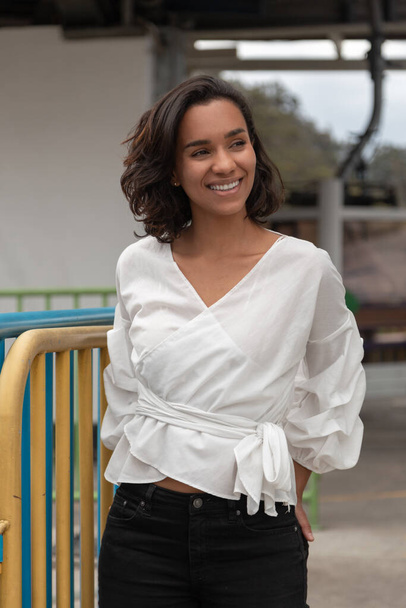 Jovem morena latina com cabelo curto e cachos, sorrindo e posando em pé, tem uma blusa branca clara e calças pretas, ao ar livre - Foto, Imagem