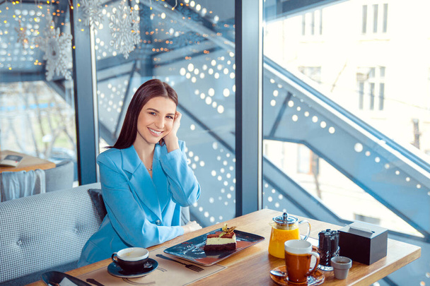Enthusiastic Γυναίκα στο μοντέρνο φθινοπωρινό ντύσιμο ψύξη σε ζεστό καφέ, προσαρμογή των μαλλιών, χαρούμενα συναισθήματα. Ταξιδιώτισσα. Ισπανίδα κοπέλα που φοράει επίσημο μπλε κοστούμι κάθεται σε ένα τραπέζι σε μια βεράντα καφέ μπαλκόνι - Φωτογραφία, εικόνα