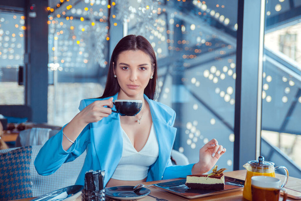 Image of young and pretty woman having dessert and drinking a coffee at home in Manhattan. Hispanic girl wearing formal blue suit sitting at table on cafe terrace balcony outside staircase background - Photo, Image