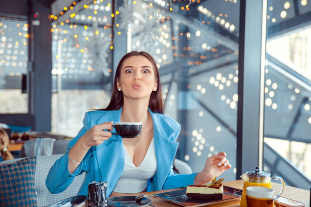 Closeup portrait of a beautiful woman puckering lips looking at camera holding cup of hot beverage. Hispanic girl wearing formal blue suit sitting at a table in a cafe or at home, near the window - Photo, image