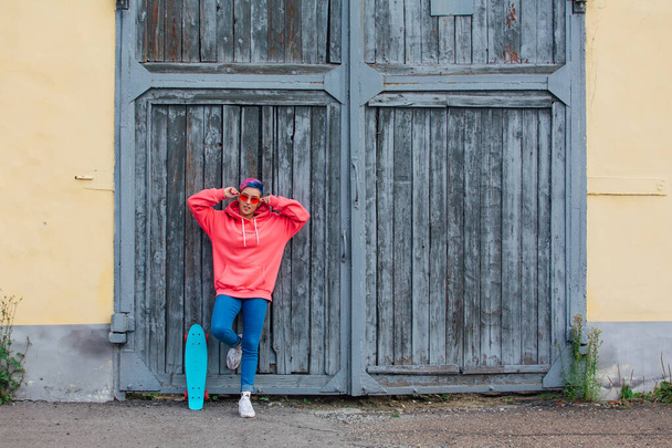Stylish young woman with short colored hair, dressed in a pink hoodie and pink glasses standing near old wooden gates with her plastic skateboard. Youth concept. - Fotoğraf, Görsel