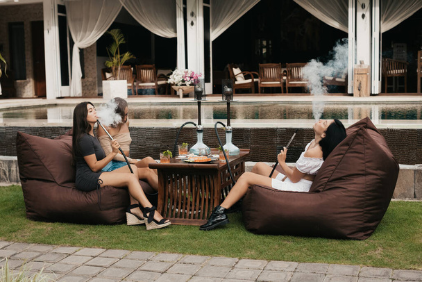Group of young women with dark hair smoking a hookah and exhaling white smoke. - Valokuva, kuva