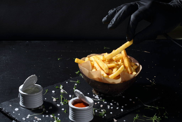 A hand in a glove takes a French fries. French fries on black background - Foto, afbeelding