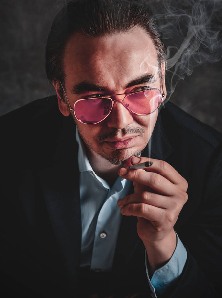 Portrait of thoughtful brutal man in official clothing and smoked sunglasses smoking cigarette and looking aside - Фото, зображення