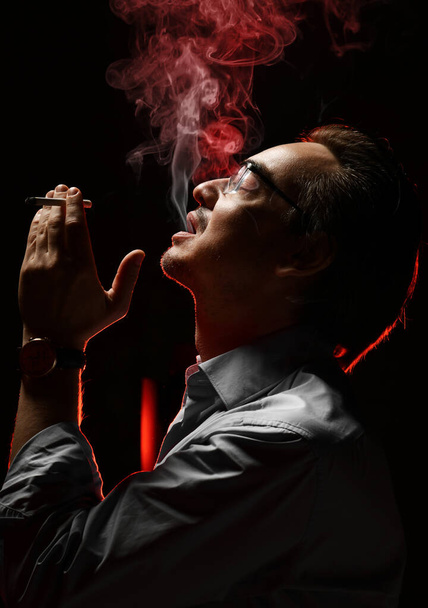 Brutaler unrasierter Mann in Diensthemd und Brille vergnügt sich beim Zigarettenrauchen und pustet Rauch aus dem Mund - Foto, Bild