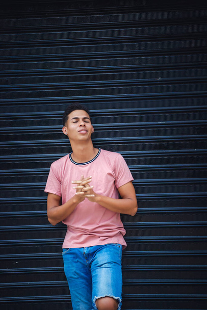 A young Hispanic male wearing jeans and a light pink t-shirt and leaning on a garage wall - Photo, image