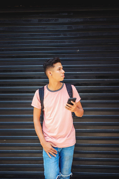 A young Hispanic male with a backpack holding a phone and leaning against a garage wall - Fotoğraf, Görsel