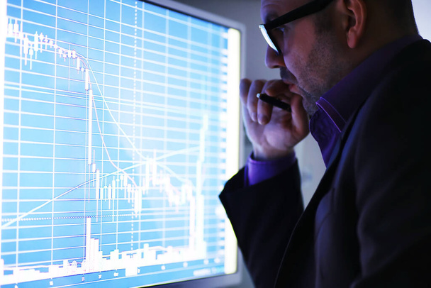 Ένας επιχειρηματίας κοιτάζει ένα γράφημα σε μια οθόνη. Ένας χρηματιστής αξιολογεί τις χρηματιστηριακές τάσεις. Ένας άνθρωπος με γυαλιά μπροστά από μια καμπύλη της δυναμικής της οικονομίας. - Φωτογραφία, εικόνα