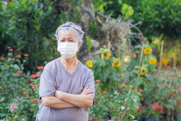 Muotokuva vanhasta naisesta, jolla oli kasvonaamio ja kädet ristissä puutarhassa. Naamio suojaamaan virusta, sepelvaltimovirusta, siitepölyä. Vanhusten ja terveydenhuollon käsite. - Valokuva, kuva