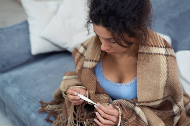 Retrato da jovem em casa com uma infecção viral. Uma mulher olha para a leitura de um termômetro. Temperatura corporal elevada. Pandemia, em auto-isolamento. - Foto, Imagem