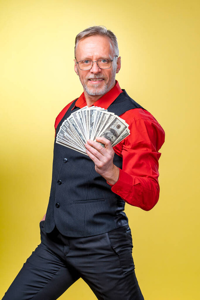 Portrait of happy and white teeth smile senior old business man holding money in hands. Dressed in red shirt. Isolated on yellow background. Human emotions and facial expressions. - Photo, Image