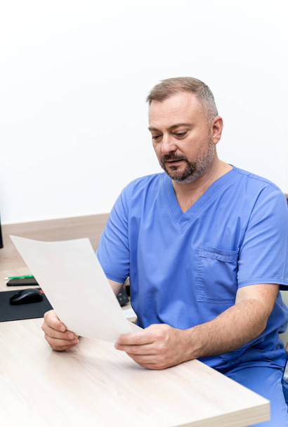 Portrait of a doctor or medical specialist. Vertical portrait. Man in scrubs sitting at table with papers in hands. - Fotoğraf, Görsel