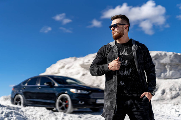 Stylish bearded young man on front view. Fashion clothes and trandy sunglasses. Luxury black car on background. White high rocks landscape. - Foto, immagini