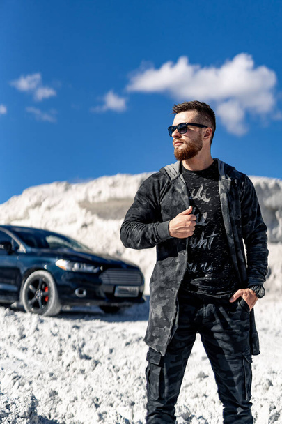 Man in cool clothes and sunglasses is posing for camera. Luxury black car on background. - Foto, imagen