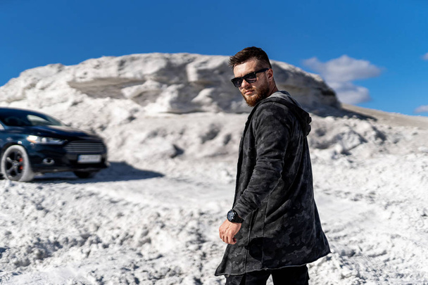 Attractive cool guy with beard stands on white rocks background. Car on white sandy road. - 写真・画像