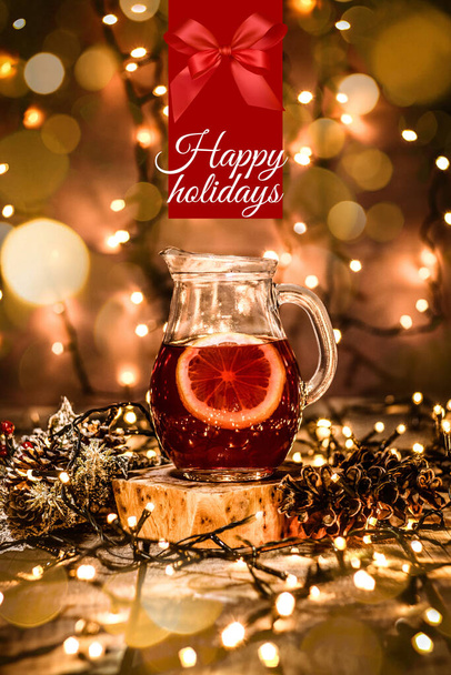 Primer plano de una jarra con té de frutas y un trozo de limón en ella sobre una mesa de madera con luces bokeh y navidad con texto Felices Fiestas - Foto, Imagen