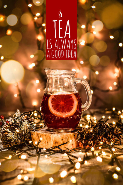 Closeup of a glass carafe with fruit tea and a piece of lemon in it on a wooden table with bokeh and christmas lights with text Tea Is Always A Good Idea - Фото, изображение