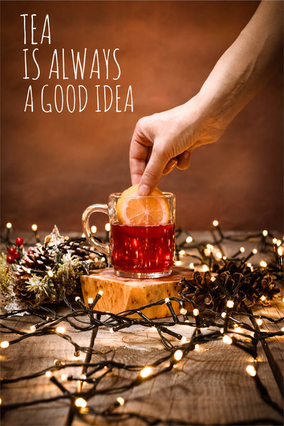 Closeup of a glass mug with fruit tea and a piece of lemon in hand on a wooden table with bokeh and christmas lights with text Tea Is Always A Good Idea - Zdjęcie, obraz