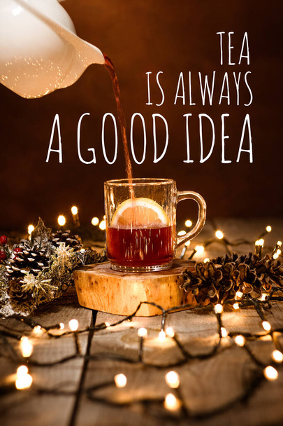 Detailní záběr skleněného hrnku s ovocným čajem a citrónem na dřevěném stole s bokeh a vánoční osvětlení s textem Tea Is Always A Good Idea - Fotografie, Obrázek