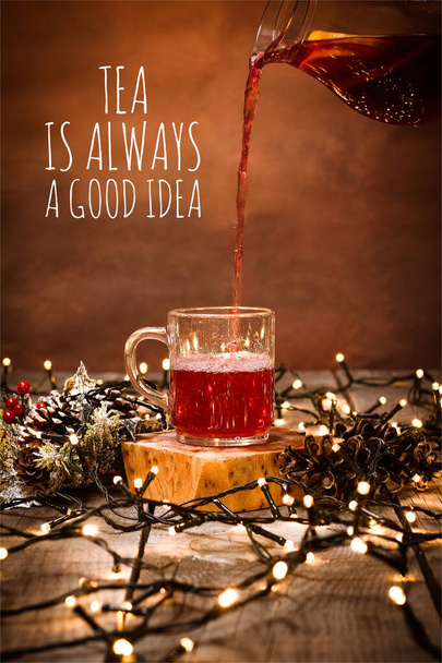 Photo of a glass mug with fruit tea and a piece of lemon in it on a wooden table with bokeh and christmas lights with text Tea Is Always A Good Idea - Foto, imagen