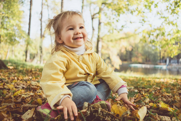 Alegre niña caucásica en otoño se sienta en las hojas en el parque. - Foto, imagen