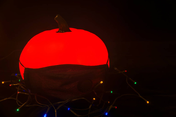 Gruselig leuchtender Terrakotta-Halloween-Kürbis ohne Augen, der eine Gesichtsmaske auf schwarzem Hintergrund mit Lichtern trägt. Halloween 2020 Feier mit COVID-19 Virenpandemie-Präventionskonzept - Foto, Bild