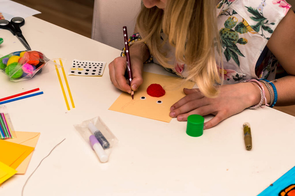 прелестная девушка с красочными бумагами, ножницами, клеем, соломинками, карандашами, лентами, кружевами на столе делает уникальные рождественские открытки - Фото, изображение