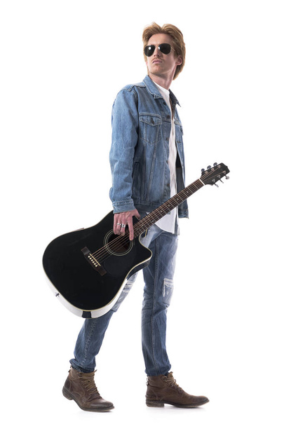Vue latérale du guitariste masculin portant la guitare marchant et regardant en arrière sur l'épaule. Corps entier isolé sur fond blanc. - Photo, image