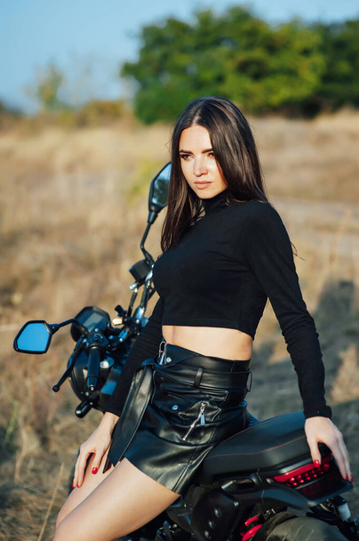 Beautiful girl in black clothes sits on motorcycle and poses for camera. Evening sunset - Foto, Imagem