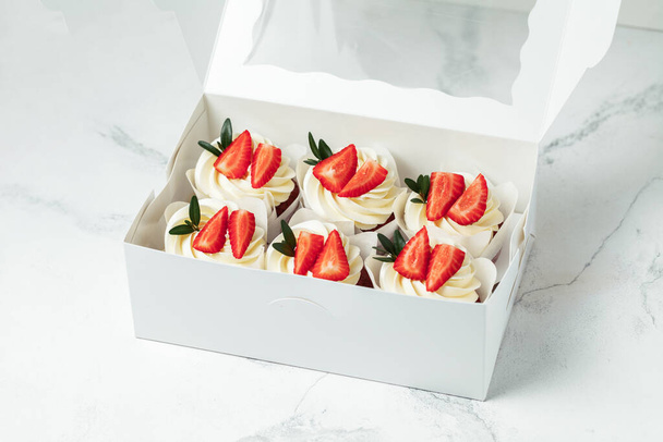 Schokoladen-Cupcakes mit weißer Sahne und Erdbeeren verziert. Muffins gestapelt in der weißen Geschenkbox auf weißem Marmorhintergrund - Foto, Bild