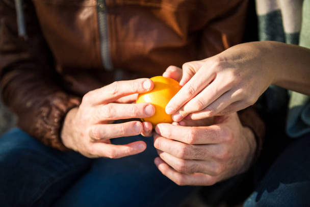 Pareja joven pela una naranja en un día soleado. Las manos humanas se acercan. amor y concepto de estilo de vida saludable. Comer vitaminas naturales al aire libre. Colaboración y trabajo en equipo en la vida real - Foto, imagen
