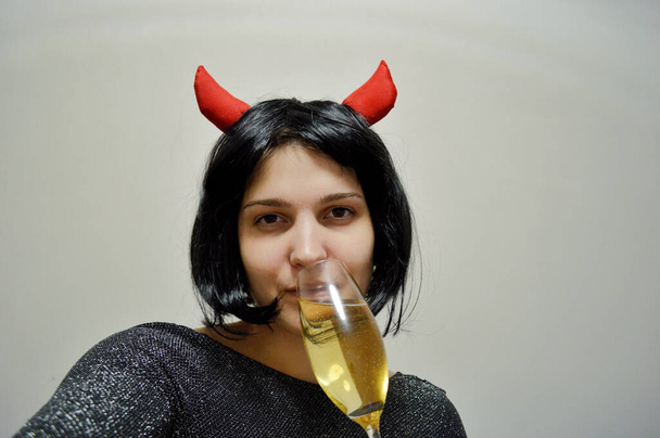 Woman with short black hair with red horns and a glass of champagne in her hand on a light background - Valokuva, kuva