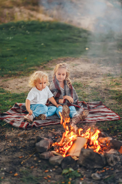 Two cute little girls sitting by a bonfire on summer evening in forest. Children are relaxing at camp bonfire. Verical portrait - 写真・画像