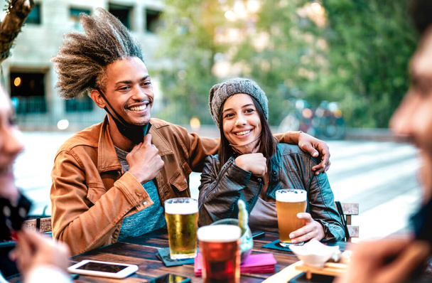 Genç çift, açık maskeli bira bardakları içiyor- arkadaşlarıyla birlikte bira barında eğlendikleri yeni normal yaşam tarzı konsepti - parlak, canlı bir filtre ve şapka takan bir kıza odaklan - Fotoğraf, Görsel