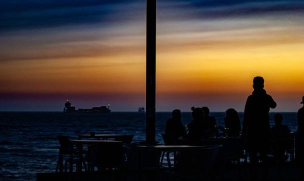 Wbrew światłu, grupa osób siedzących na tarasie baru delektuje się niesamowitym zachodem słońca nad morzem w Porto w Portugalii. Jeden z tych ludzi stoi. W tle, w morzu, można zobaczyć kilka łodzi - Zdjęcie, obraz