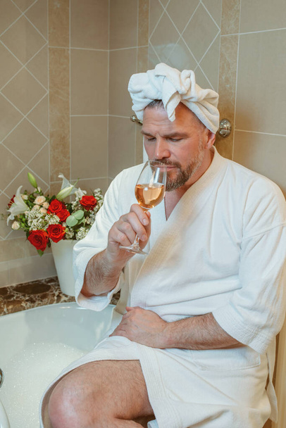 knappe volwassen man in witte badjas en handdoek op zijn hoofd zittend op badkamer met schuim en het drinken van rozenwijn. Spa, ontspannen, levensstijl, genieten van het leven concept. - Foto, afbeelding