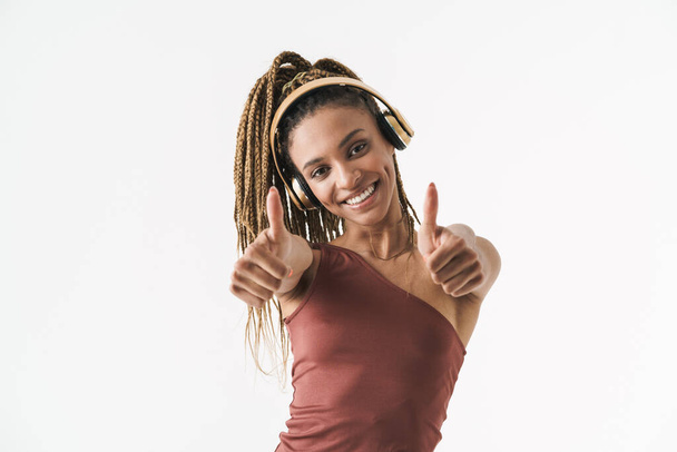 Retrato de bela mulher africana morena sorridente com dreadlocks uma música ouvindo via fones de ouvido sem fio isolados em fundo branco, polegares para cima - Foto, Imagem