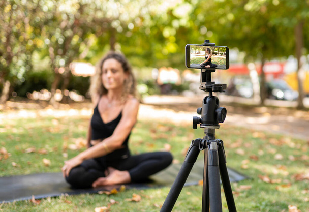 Femme assise sur le tapis enseigne le yoga dans le parc à travers un smartphone sur trépied, éducation en ligne. - Photo, image
