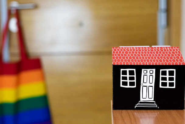 Ön planda küçük bir oyuncak ev görebilirsiniz, ve arka planda, bir evin dış kapısı, ve tutamağa asılı eşcinsel gurur bayrağı renginde bir çanta veya LGBTİ - Fotoğraf, Görsel