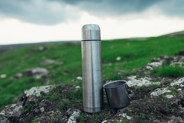 Camping and travel concept. Close-up of steel thermos and stainless cup, on stone in green field of mountains. - Photo, Image